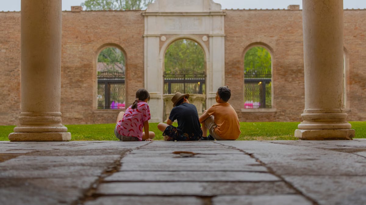 Three kids sit together in an ancient cloister, surrounded by marble pillars and lush greenery.